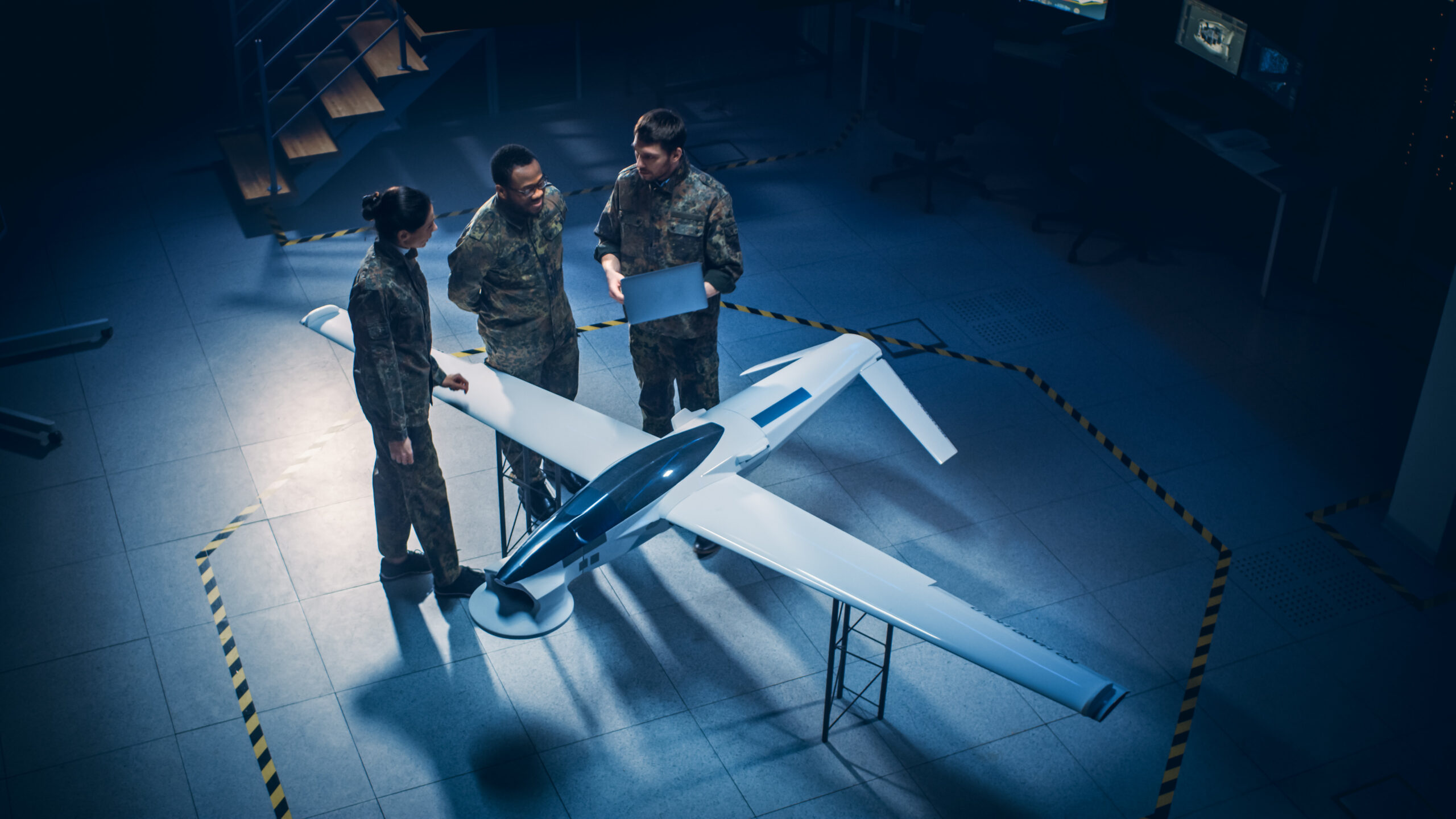 Army Aerospace Engineers Work On Unmanned Aerial Vehicle / Drone. Uniformed Aviation Experts Talk, Using Laptop. Industrial Facility with Aircraft for Performing Surveillance, Warfare Tactics, Attack